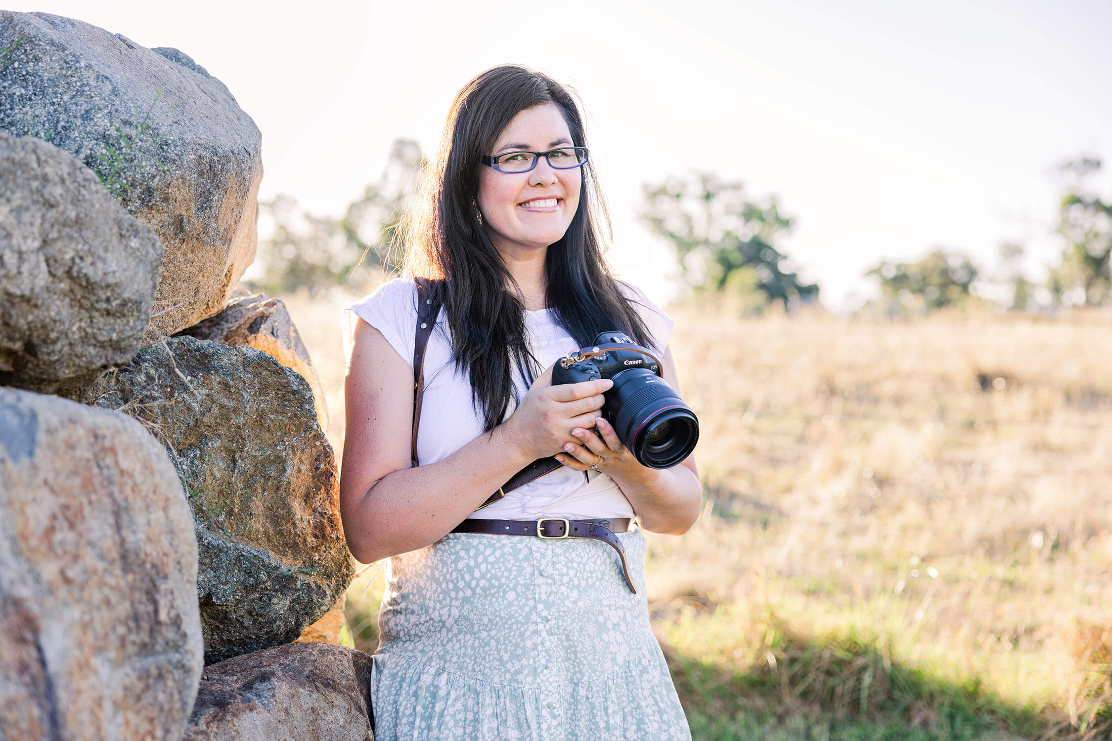 photographer with long dark hair standing in front of rocks with a sunset in the background