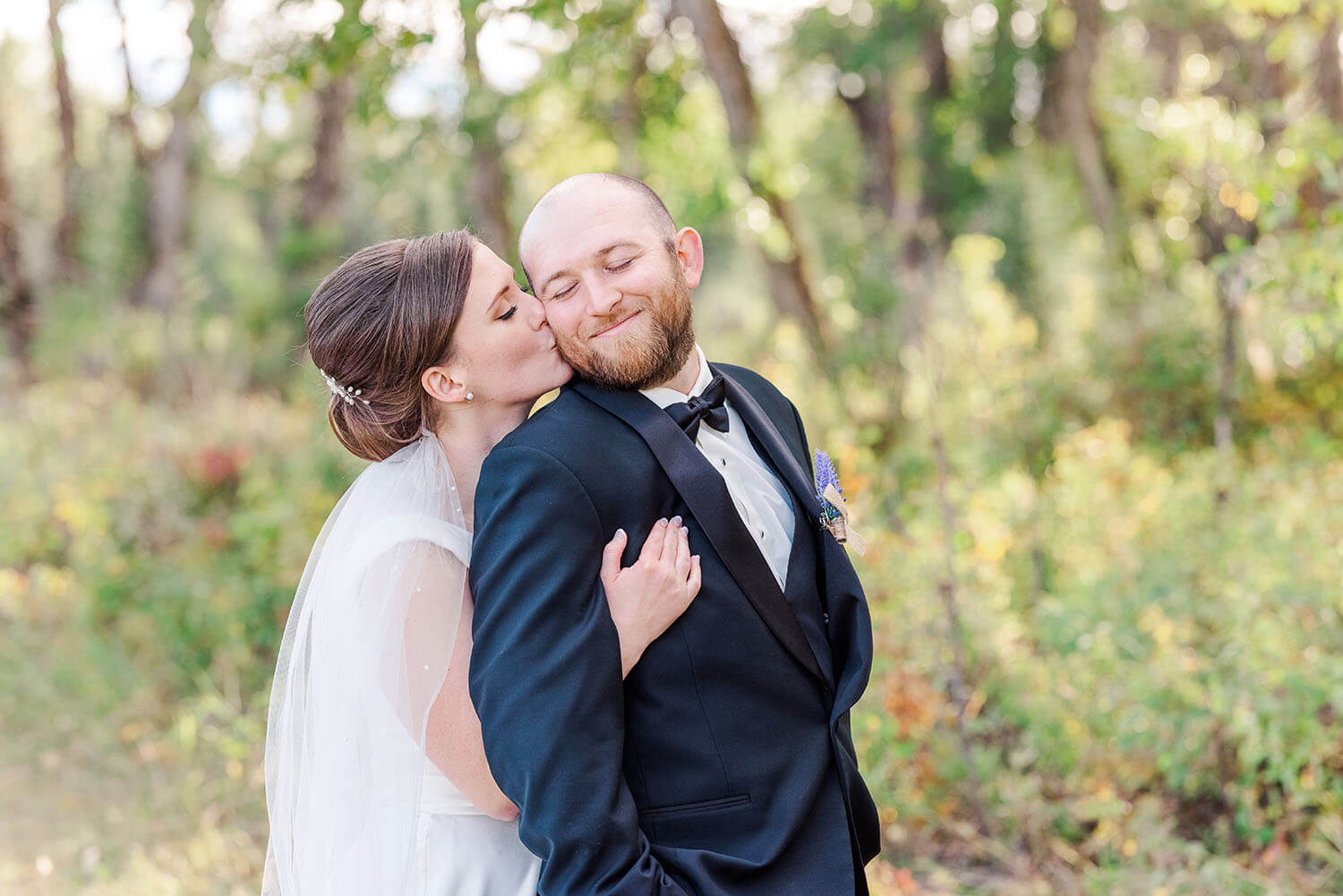 a bride standing behind her groom giving him a big kiss on the cheek while hugging him