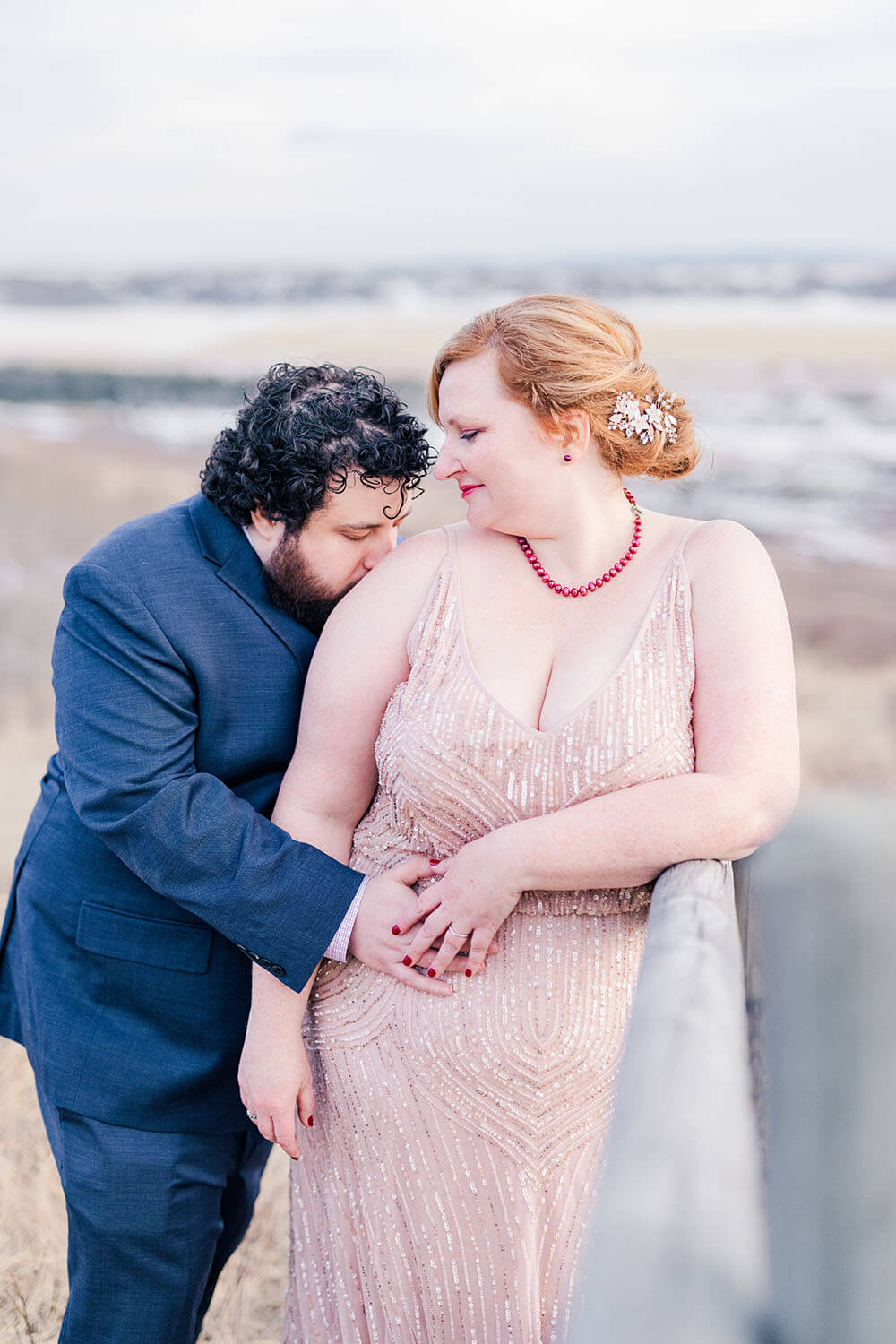 groom in navy blue suit with dark curly hair kissing the shoulder of his red haired bride in a blush pink beaded wedding dress