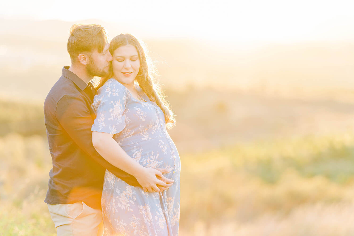 sunset behind a pregnant woman and her partner is kissing her on the cheek