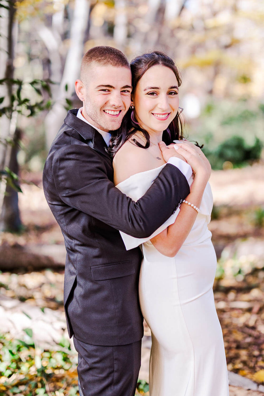 groom in black suit hugging his bride from behind with autumn forest behind them