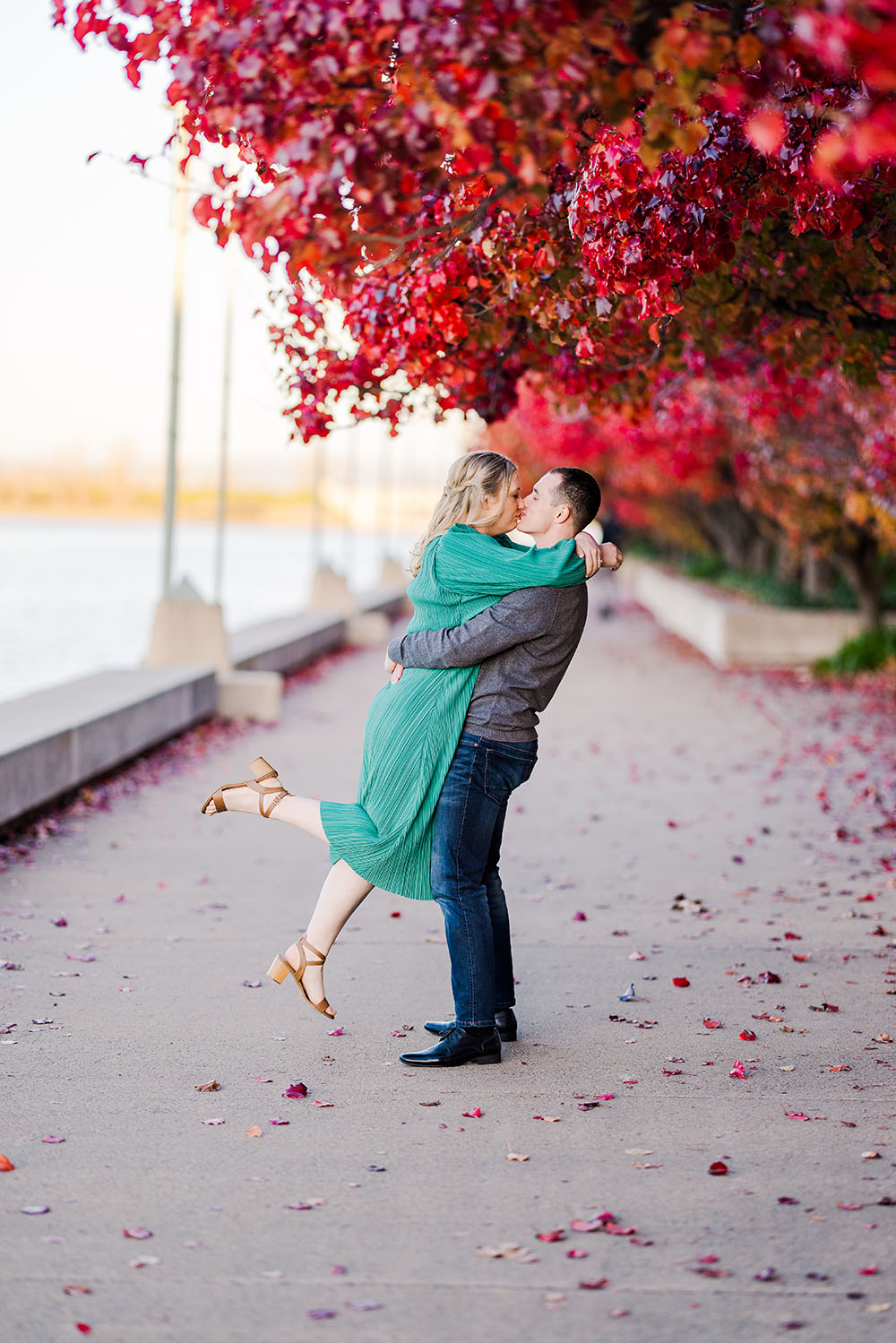 man lifting a blonde woman in a green dress under a tree with red leaves