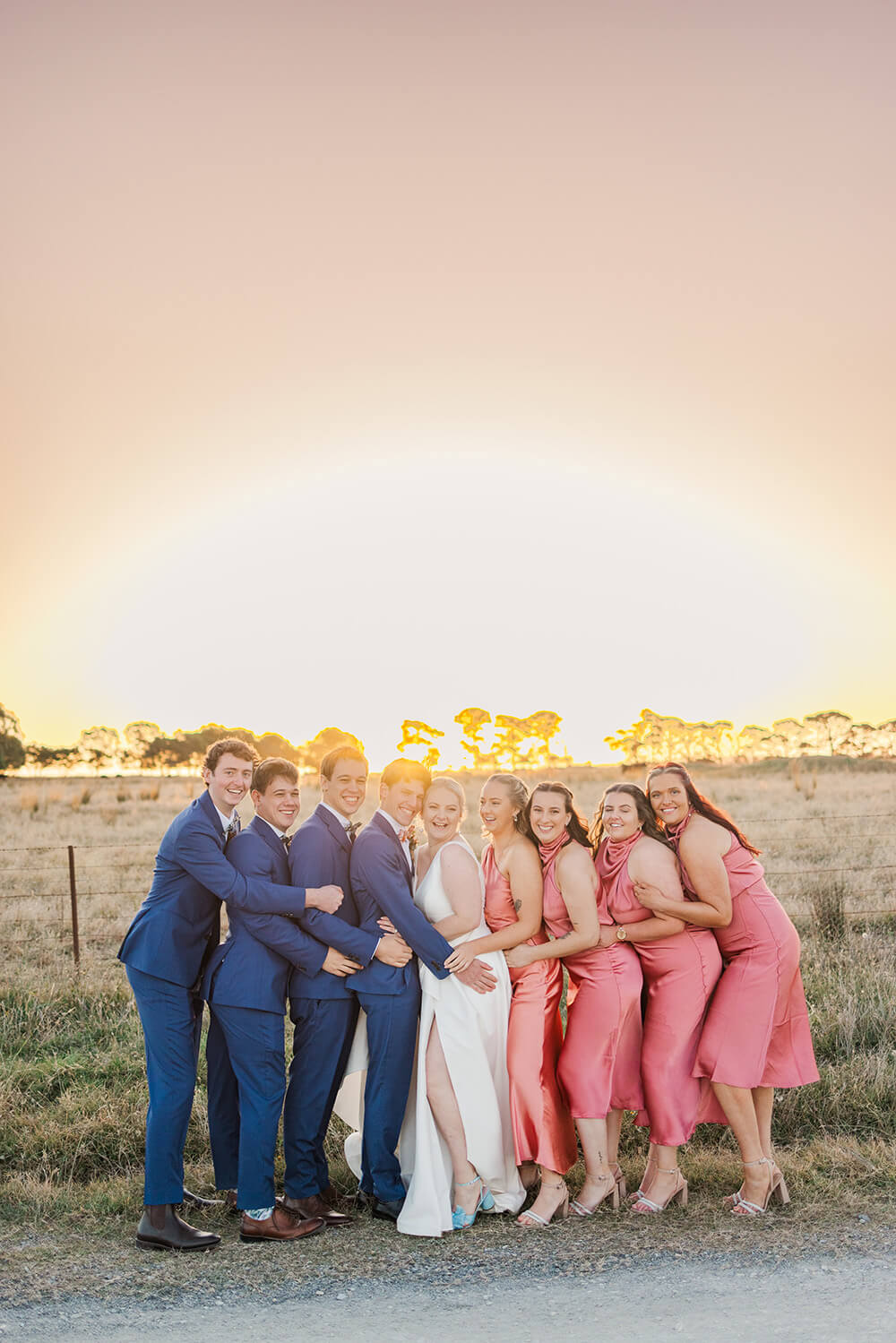 4 groomsen in navy blue suits and 4 bridesmaids in coral pink squeezing a bride and groom and smiling for a photo