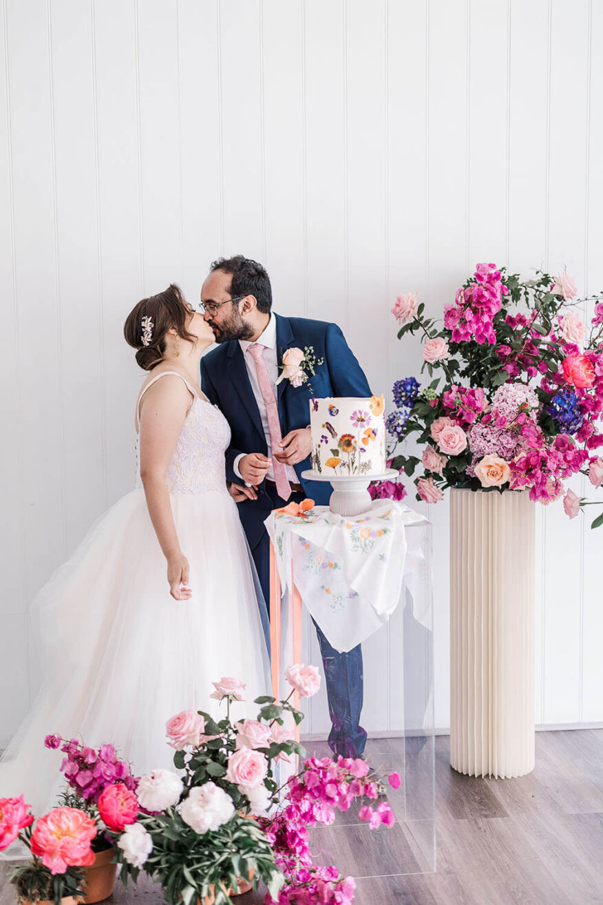 Bride and groom having a kiss over the top of a flower adored wedding cake