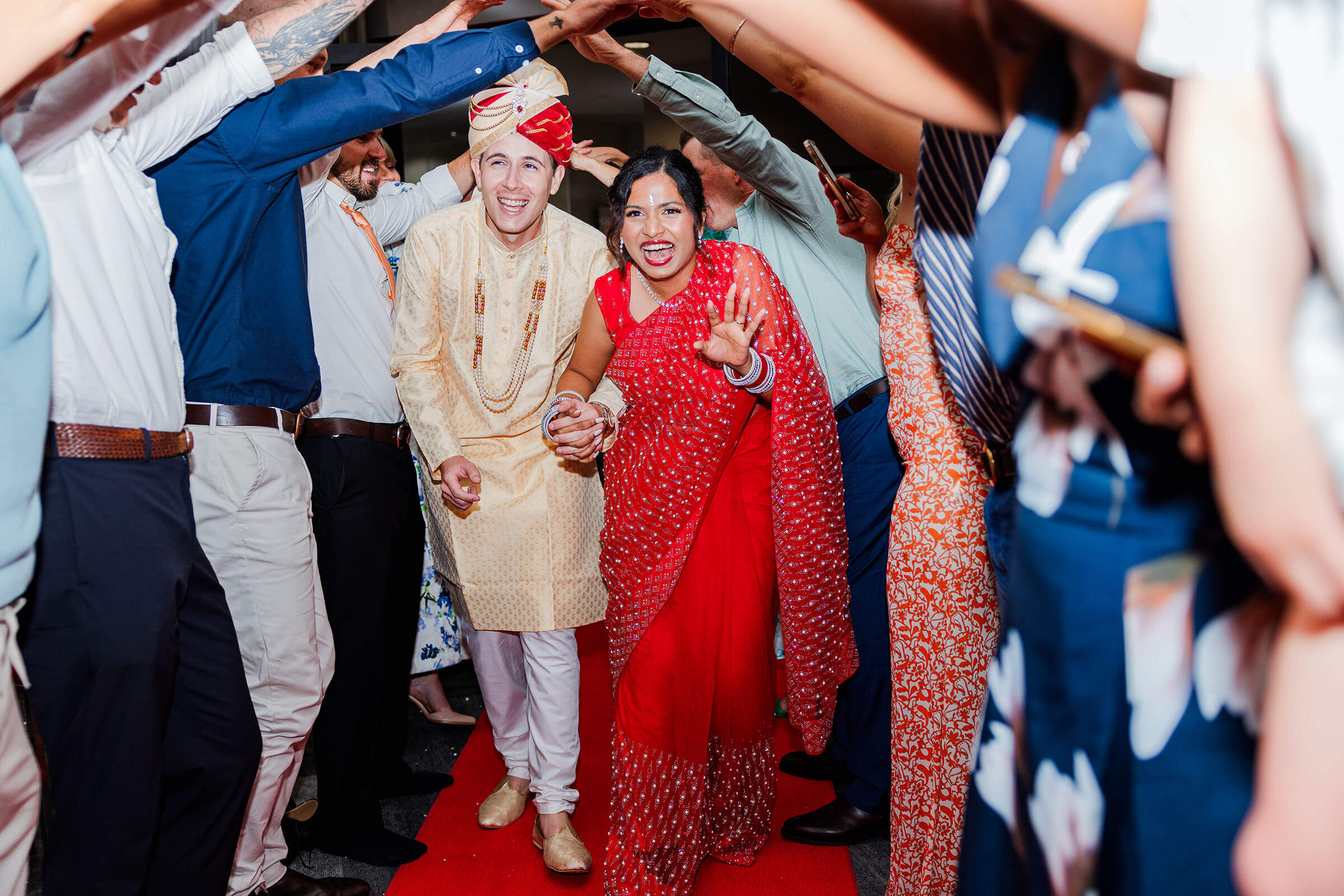Indian bride in a red sari holding hands with a causian groom in traditional golden indian outfit running through a tunnel formed by guests
