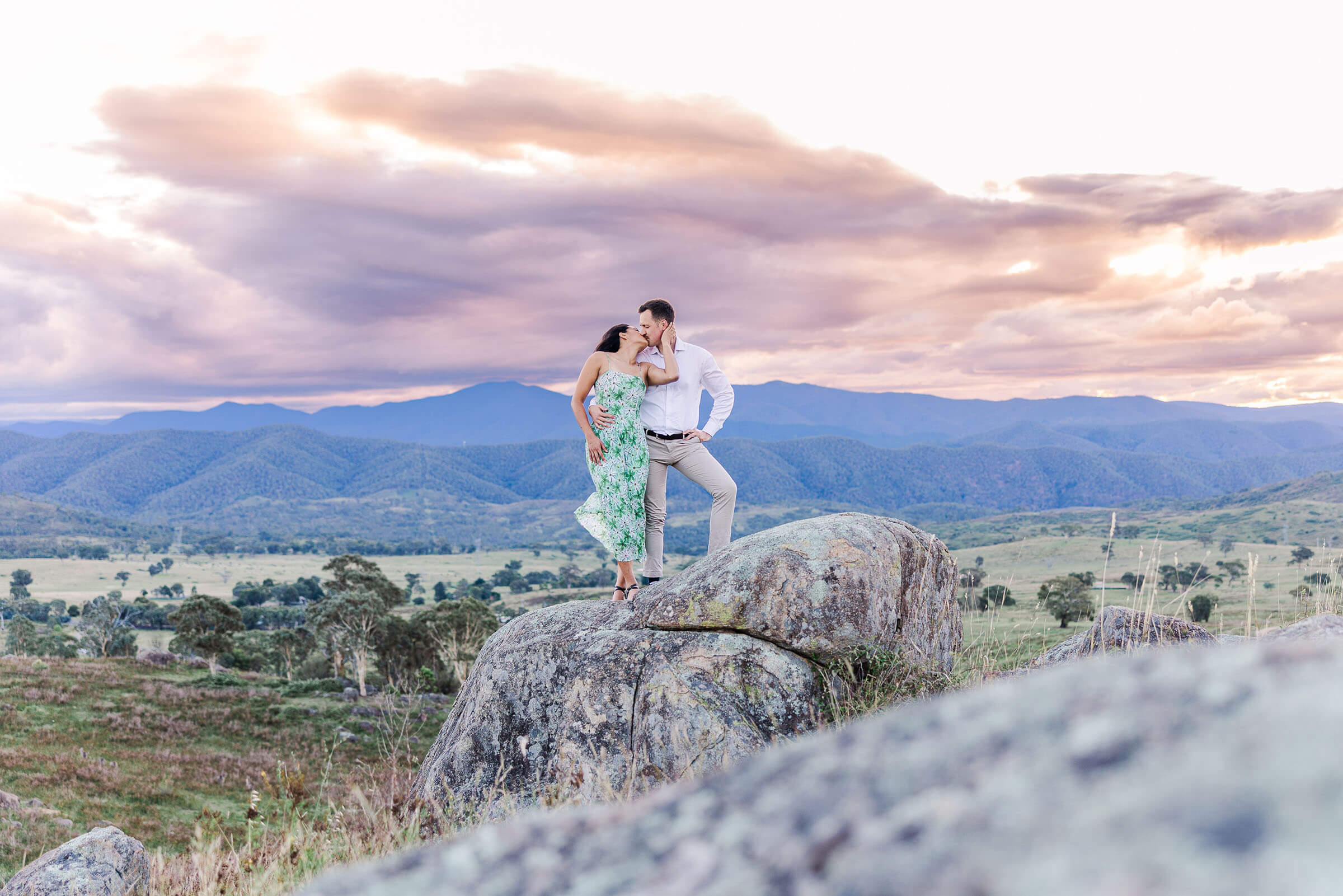 an Indian woman in a green dress being kissed by a causain man in a white shirt, standing on a boulder with a sunset behind