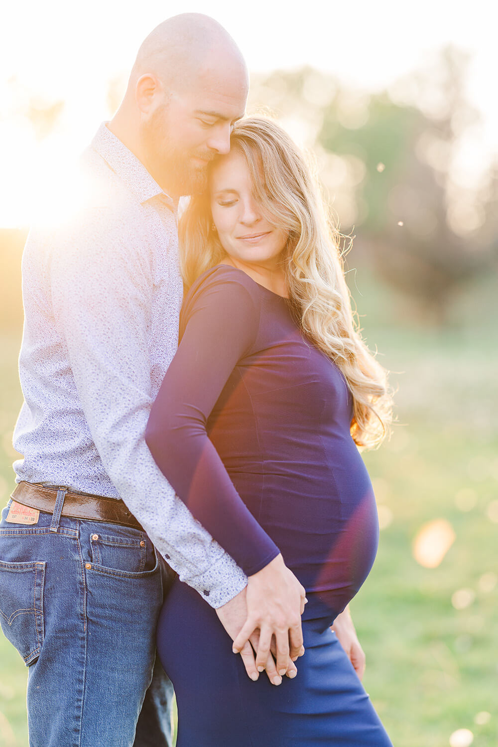 man hugging his pregnant wife from behind with sun shinning behind them