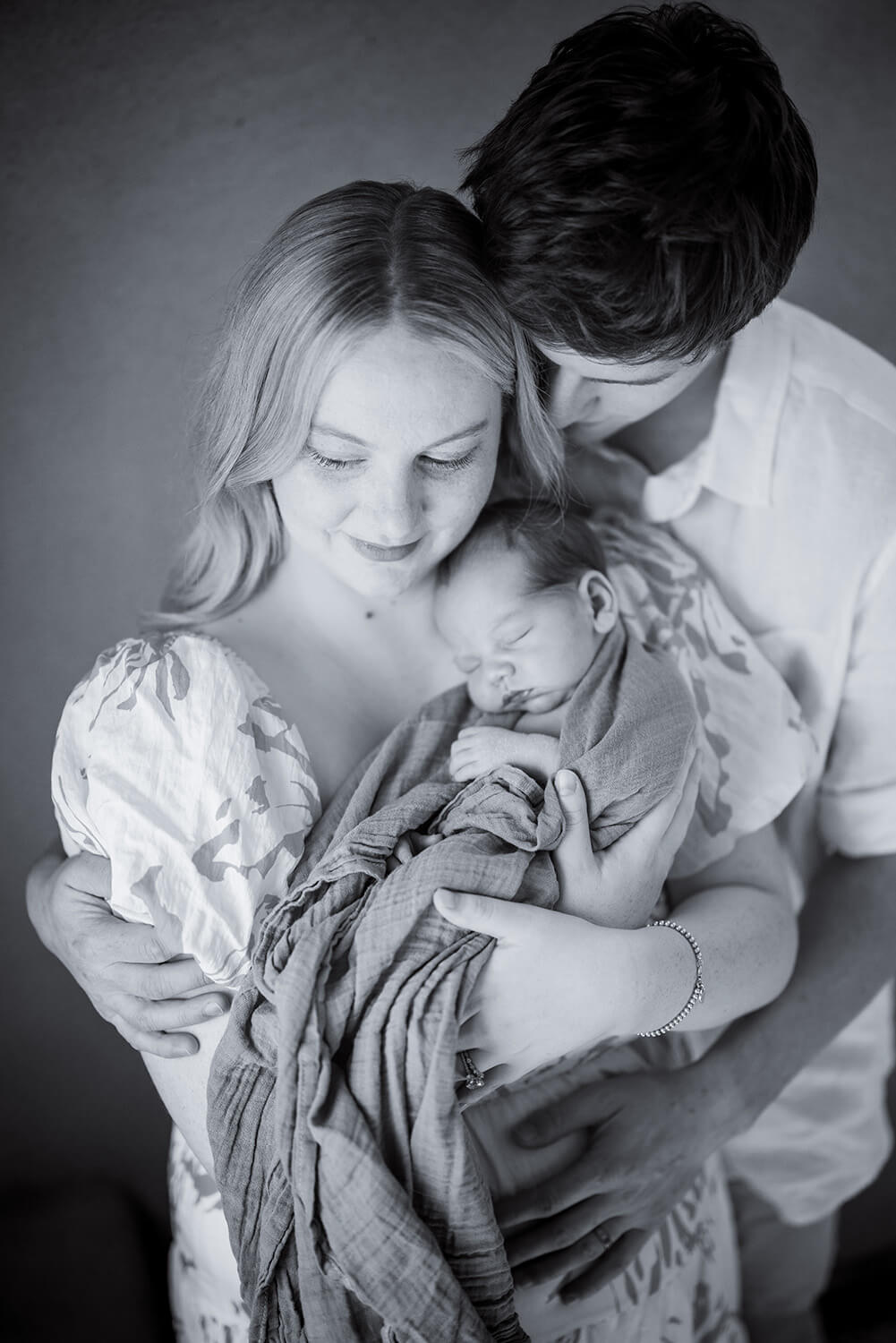 mother holding newborn baby against her chest with the father hugs both of them from behind