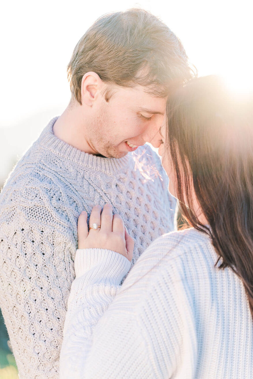 a brunette couple hugging with foreheads together. The woman has an engagement ring on her hand place on the mans chest.